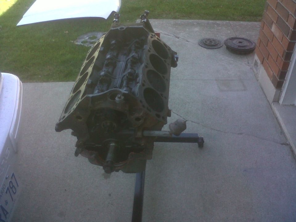 Engine block on a stand, with a white car part and a brick wall in the background