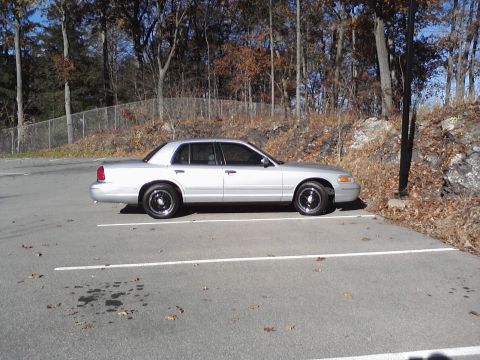 2000 Ford Crown Victoria - A silver 2000 Ford Crown Victoria is parked in an outdoor lot with trees in the background.