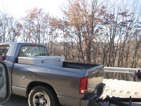 1991 Ford Bronco - A gray pickup truck is parked on the side of a road, with a toolbox secured in its bed and trees visible in the distance.