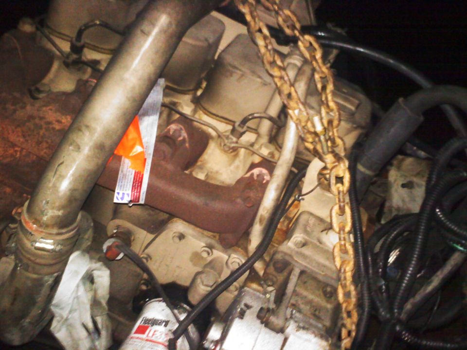 Engine block with exhaust manifold, oil filter, and lifting chains. Orange tag attached to manifold