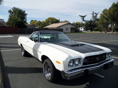 1973 Ford Ranchero - A white 1973 Ford Ranchero with a black hood stripe and vinyl top is parked in an empty lot.