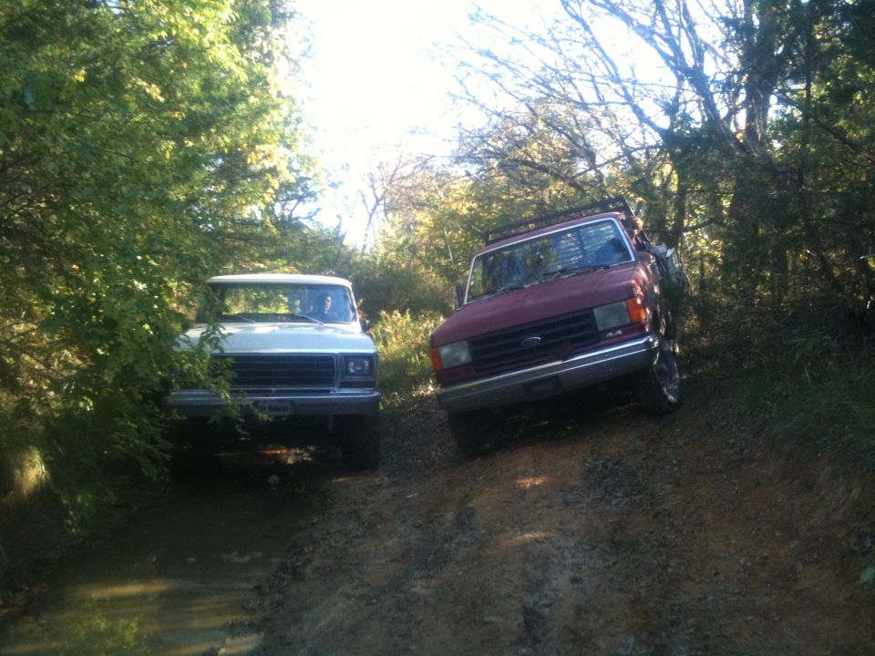 Two trucks, one white and one maroon, are on a muddy dirt path surrounded by trees. The maroon truck has a roof rack
