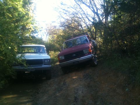 1978 Ford Bronco - Two trucks navigate a muddy trail through a wooded area.