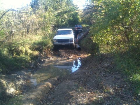 1978 Ford Bronco - Two vehicles navigate a challenging muddy terrain with trees lining the path.
