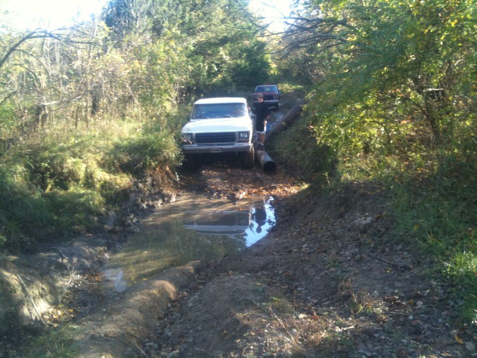 White 1978 Ford Bronco driving through a muddy ditch with a second vehicle behind it
