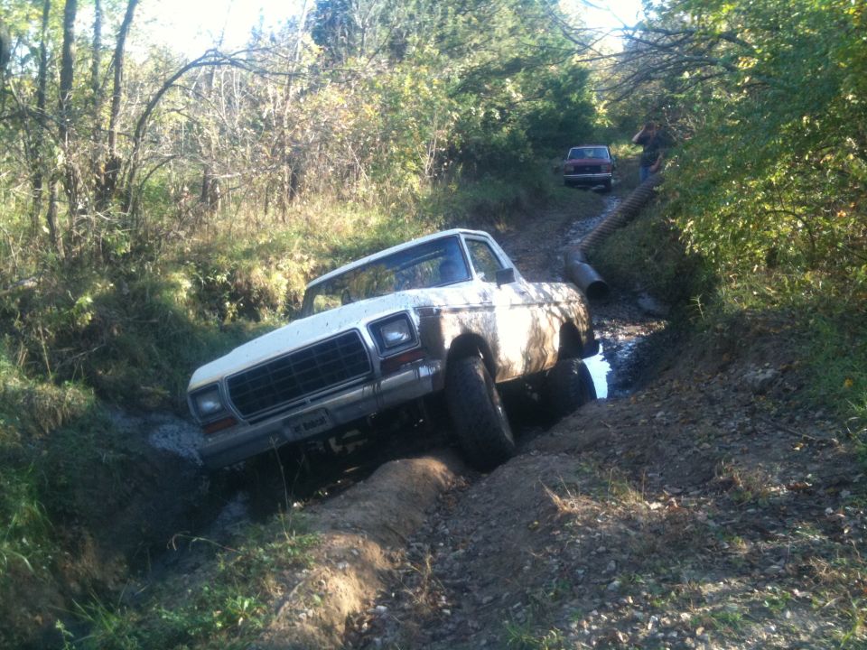 White pickup truck stuck in mud on a trail. Another red vehicle is further up the trail