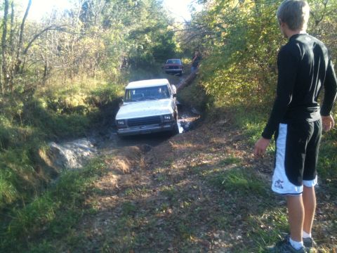 1978 Ford Bronco - A white SUV attempts to drive through a muddy path, with a second vehicle visible further down the trail.