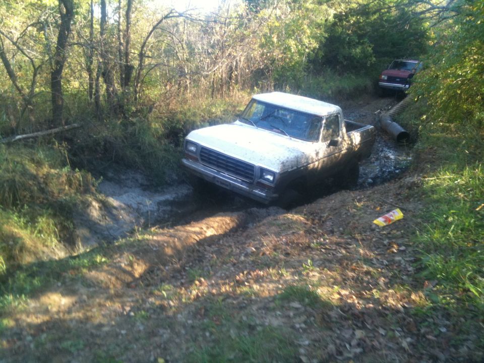 White Ford pickup truck stuck in a muddy ditch, another red vehicle behind it