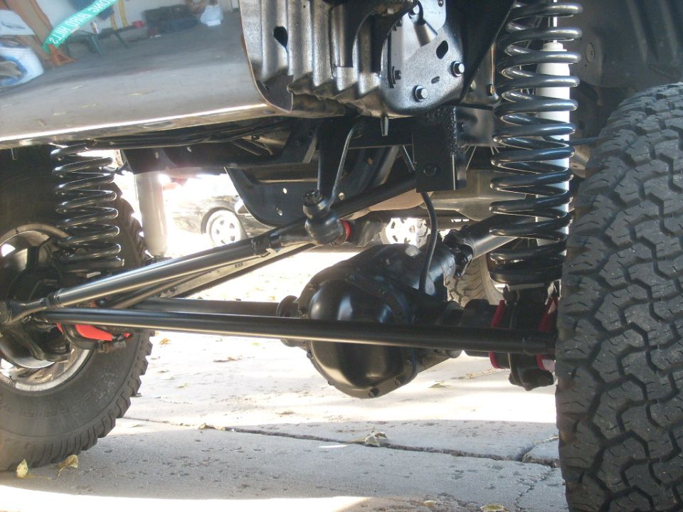 Underside of a vehicle showing suspension, differential, and tire. Another car is visible in the background
