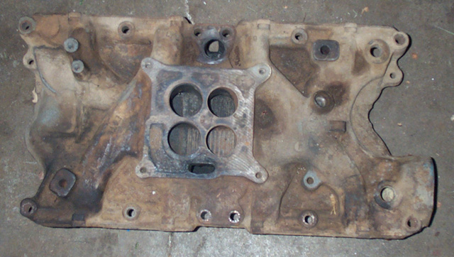 Rusty cast-iron V8 intake manifold with square-bore four-barrel carb mount on concrete