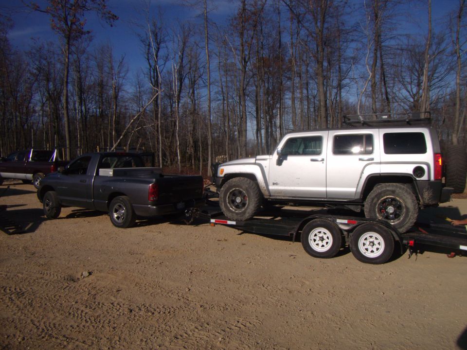 Silver Hummer H3 on a trailer being towed by a gray Dodge Ram pickup. Trees in background