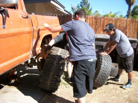 1969 Ford F-250 - Two men help a third man remove a large tire from an orange truck that is jacked up.