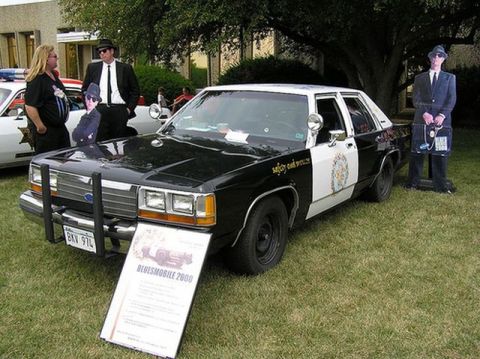 1991 Ford Crown Victoria - A 1991 Ford Crown Victoria, modified as the Bluesmobile, is displayed with costumed individuals and informational signage.