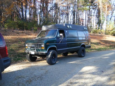 1981 Ford E350 - A modified van with a raised suspension and large tires sits parked on a dirt road near a wooded area.