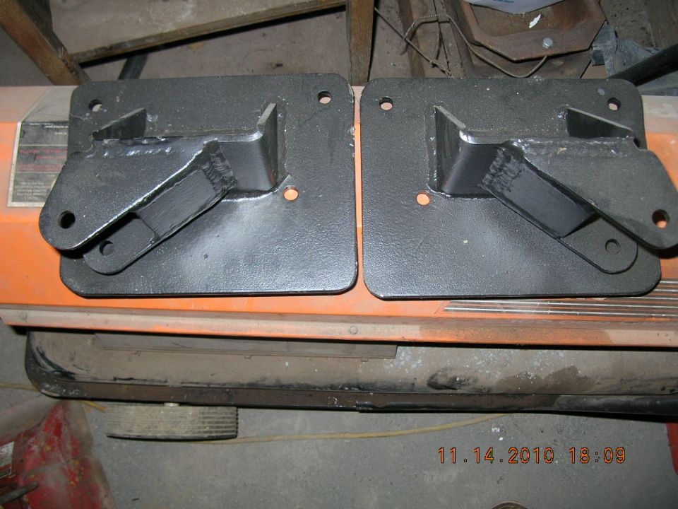 Two black metal brackets with welded supports and mounting holes, resting on an orange surface