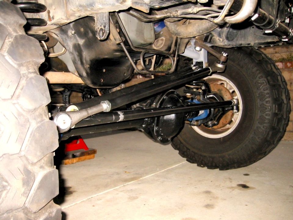 Underside of lifted 4x4 front axle with black diff, steering linkage, and mud tire in garage