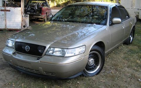 2000 Mercury Grand Marquis - A gold Mercury Grand Marquis is parked in a grassy area next to a trailer and a red pickup truck.