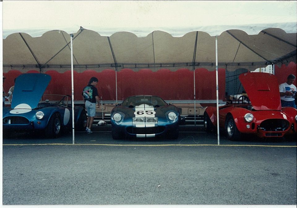 Blue and red Cobra roadsters flanking a numbered 65 blue race coupe under a paddock tent