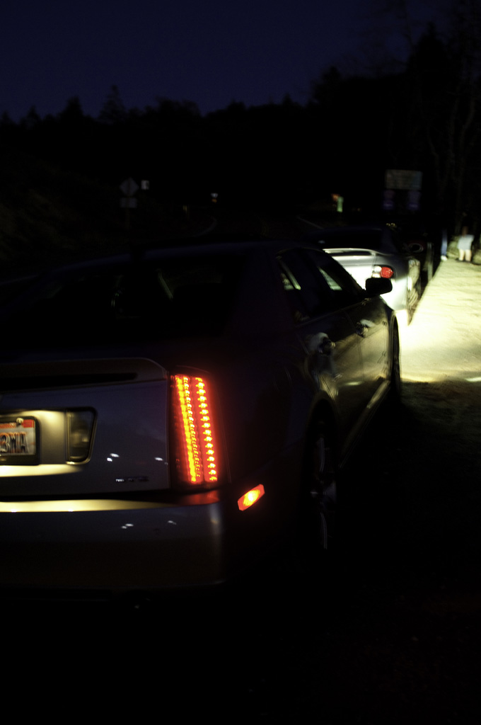 Cadillac STS rear with illuminated taillights and license plate visible. Another car is parked behind it