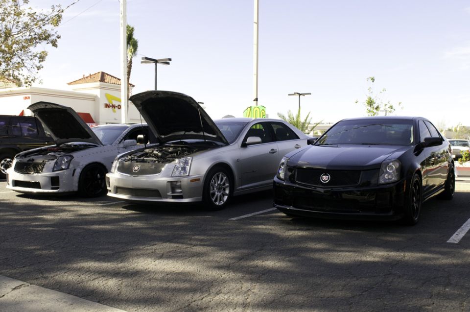 Three Cadillac sedans parked in a lot, two with hoods open. Black Cadillac in foreground