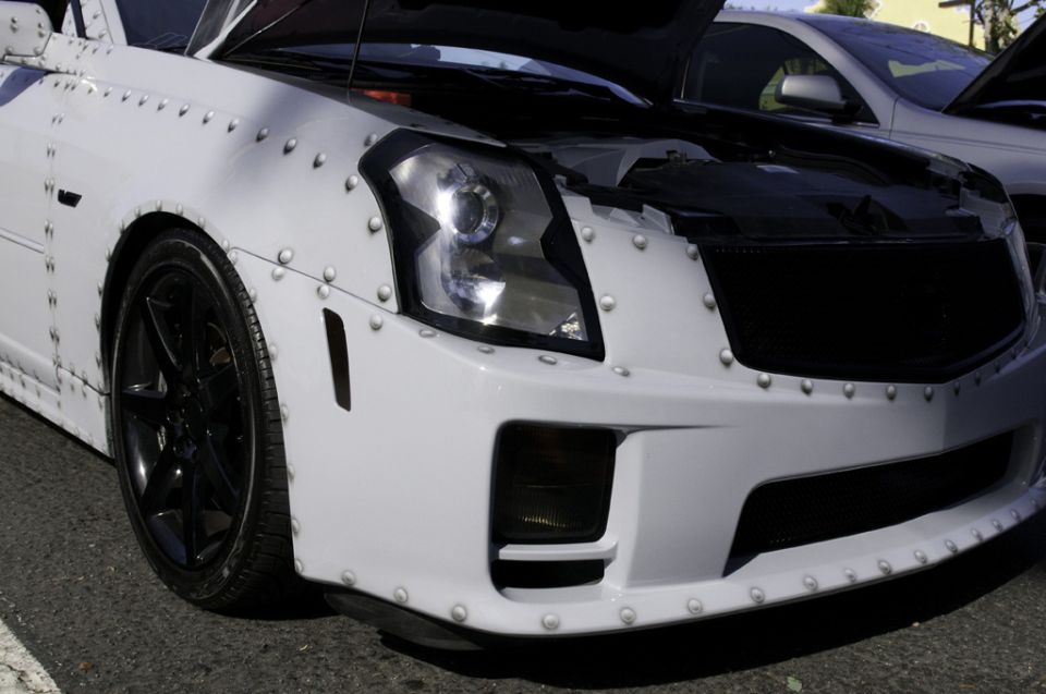 White car with rivets all over its body panels, hood open, parked next to another car