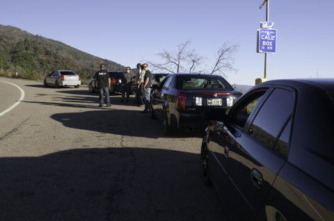 2006 Cadillac STS - A group of people gathered around several Cadillac sedans parked on a scenic roadside with a call box nearby.