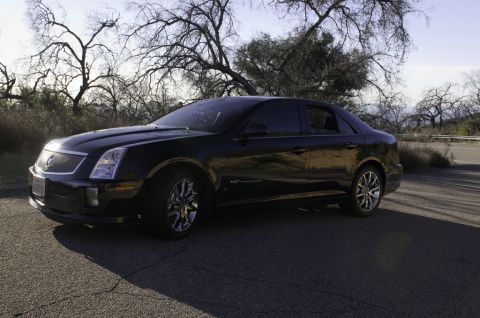 2006 Cadillac STS - A black 2006 Cadillac STS is parked on a cracked asphalt road with bare trees in the background.