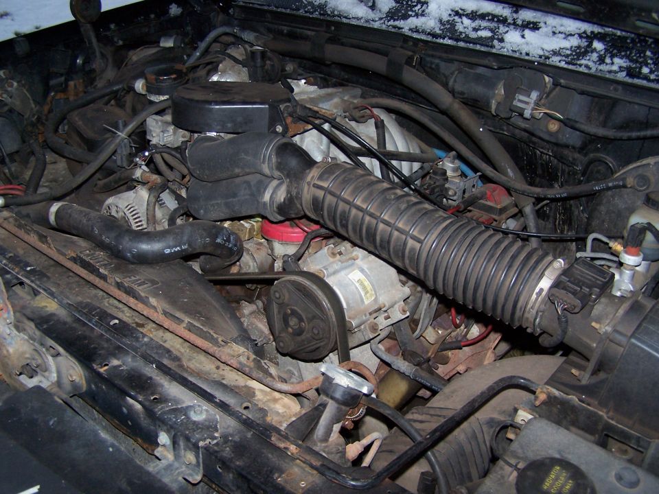 Engine bay with hoses, belts, and engine components visible. Some snow on the upper edge