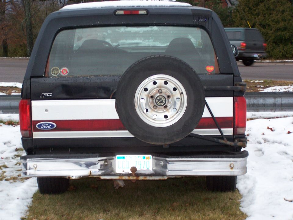 Rear view of a black Ford Bronco with a spare tire mounted on the back. Another SUV is visible in the background