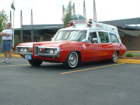 1999 Ford Crown Victoria - This vintage ambulance, likely a 1969 Pontiac, is parked outside a building with a man standing nearby.