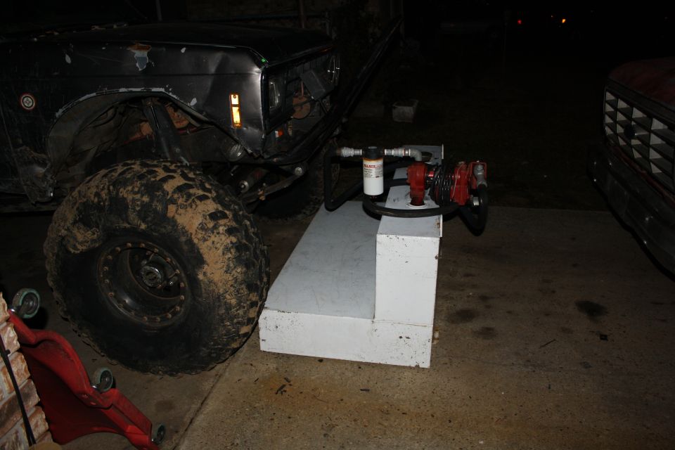 Muddy off-road vehicle with a large tire, next to a white fuel tank and pump. Another truck is partially visible on the right