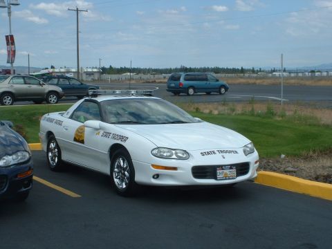 1999 Ford Crown Victoria - A white Chevrolet Camaro, marked as a State Trooper vehicle, is parked in a lot with other cars visible.
