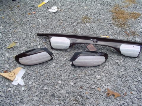 1999 Ford Taurus - Car parts, likely side mirrors and a rear trim piece, are laid out on a gravel surface, some covered with tape.