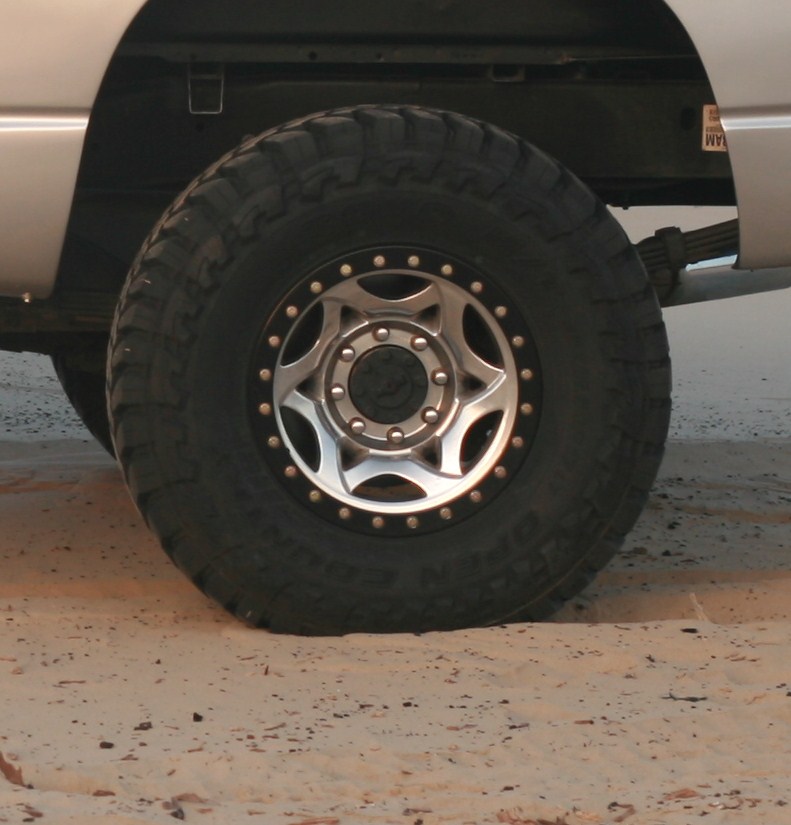 Close-up of a large off-road tire and custom rim on a silver truck, parked on sand