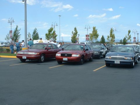 1999 Ford Crown Victoria - A red 1999 Ford Crown Victoria is parked in a lot with other vehicles under a bright sky.