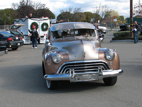 Front view of a two-tone brown and tan vintage car with chrome grille and bumper. Other vehicles and people are visible in the background