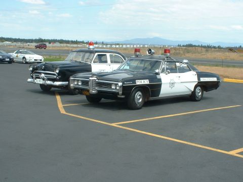 1999 Ford Crown Victoria - Two vintage police cars, one a Pontiac, are parked in a lot under a partly cloudy sky.