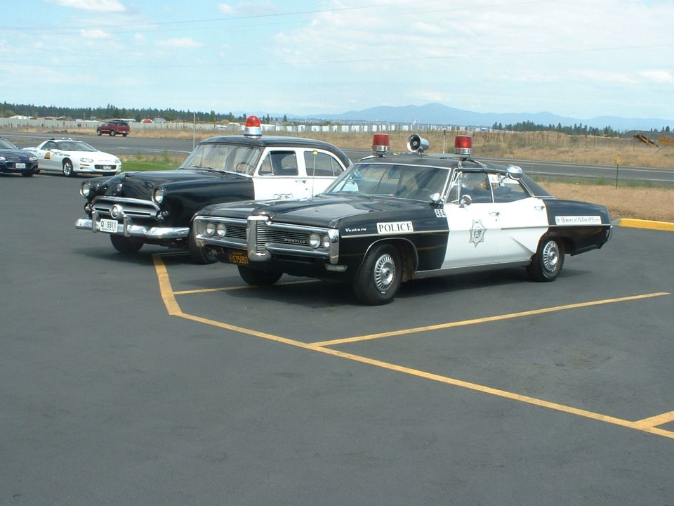 Black and white police car with 'POLICE' on door, parked next to older black and white police car