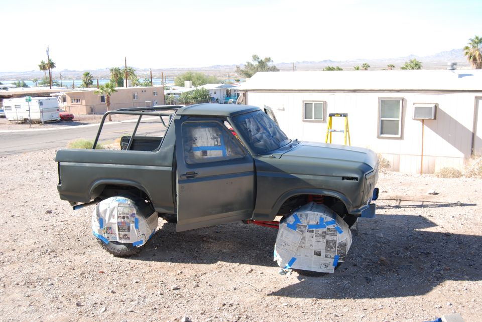 1980 Ford Bronco with doors open, wheels taped off for painting. Ladder and trailer in background