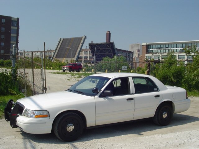 2003 Ford Crown Victoria police car parked on dirt lot with industrial buildings and a drawbridge in background