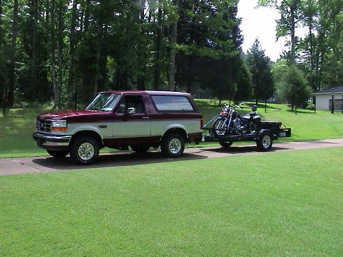 Dark red and tan Ford Bronco towing a trailer with a motorcycle on a driveway among trees