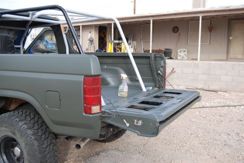 1982 Ford Bronco - Rear view of a gray Ford Bronco with its tailgate open, revealing a roll cage and cleaning supplies in the bed.