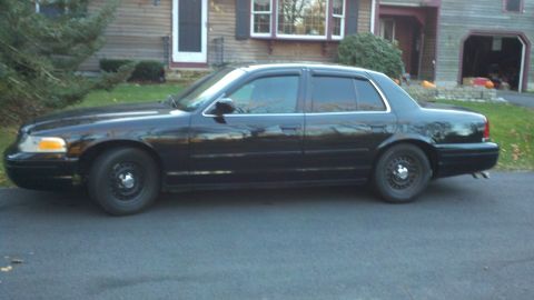 1999 Ford Crown Victoria - Black sedan with tinted windows and steel wheels parked on a paved surface in front of a residential building.