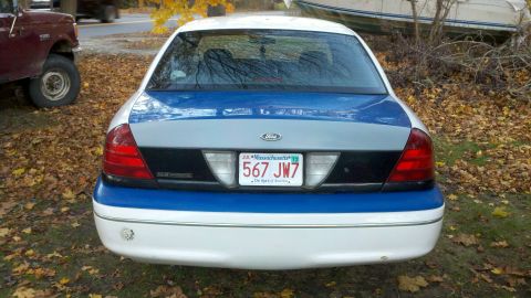 2003 Ford Crown Victoria - A 2003 Ford Crown Victoria is parked next to a red truck, with fallen leaves on the ground.