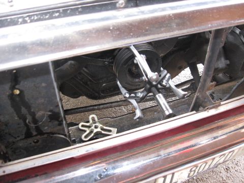 1981 Ford Bronco - View through the chrome grille opening of a dark red truck with the radiator removed, showing a jaw-type puller clamped onto the multi-groo…