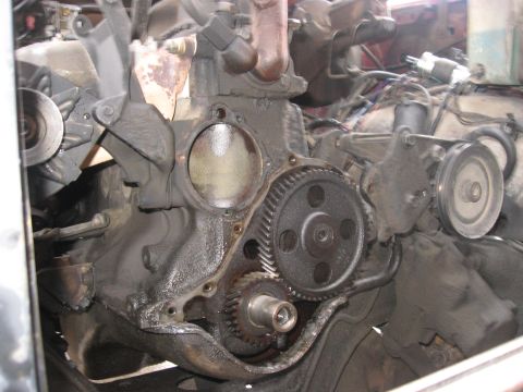 1981 Ford Bronco - Front of a V8 engine with the timing cover and water pump removed in the vehicle, exposing the large helical-cut camshaft gear meshed with…