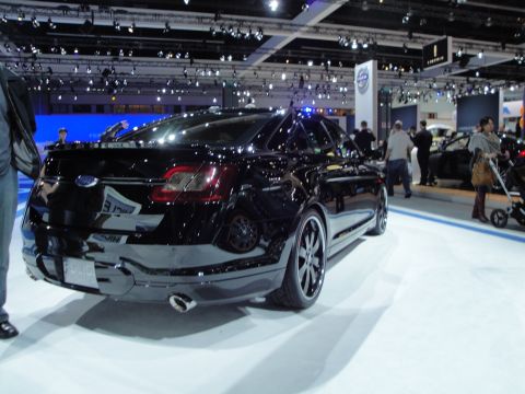 1993 Ford Taurus - A black sedan is displayed at an auto show, with people and other vehicles in the background.