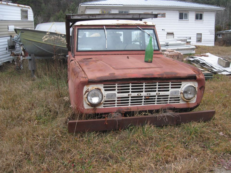 Rusty red Ford Bronco with a white grille and a green bottle on the hood. Trailer and boat in background