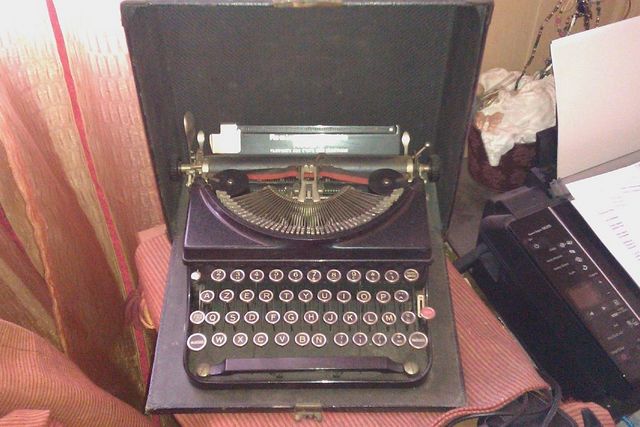 Black typewriter with a metal carriage and keys, inside an open case. Text visible on the carriage