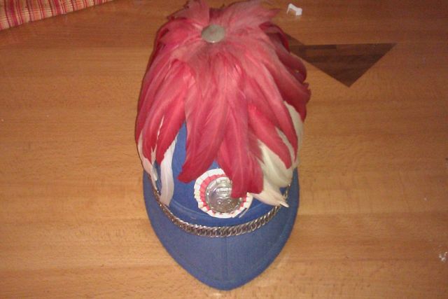 Blue hat with red and white feathers on top, silver chain across brim, and a circular emblem